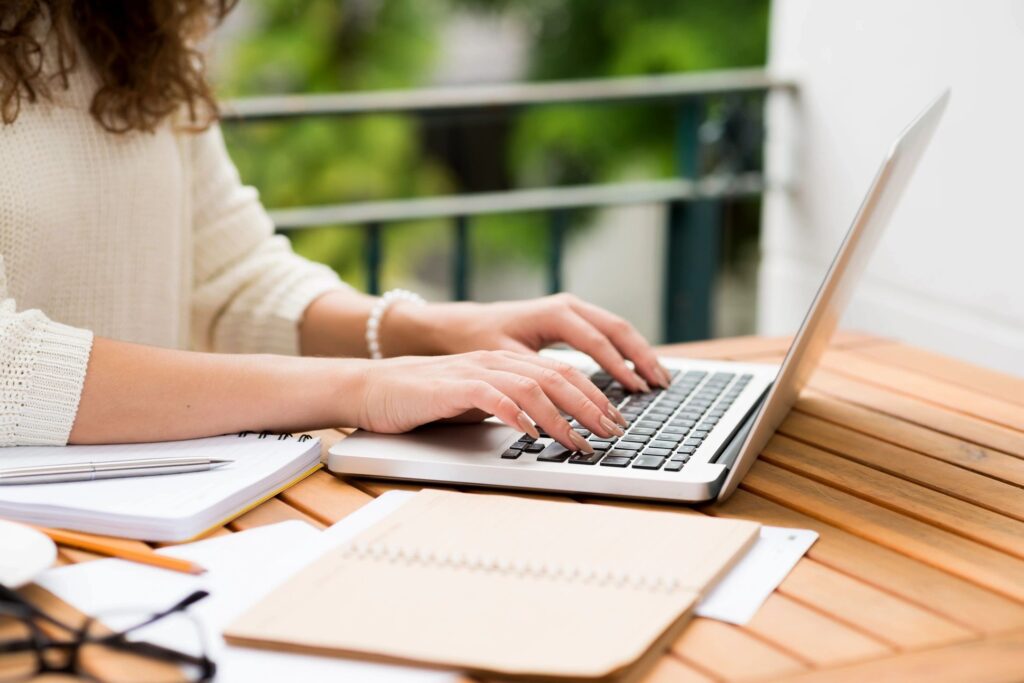 Person typing on a laptop outdoors with notebook and pen nearby.
