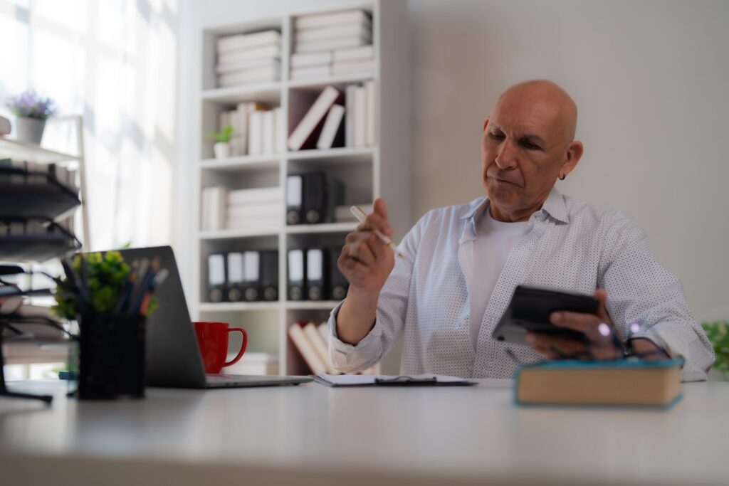 An older man in a light shirt works with a calculator and notebook beside a laptop at a bright desk