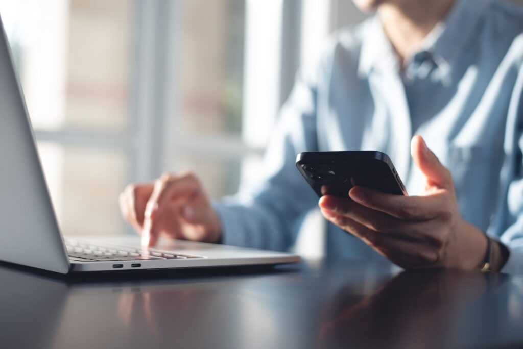 Close-up of a person using a smartphone in one hand while typing on a laptop with the other