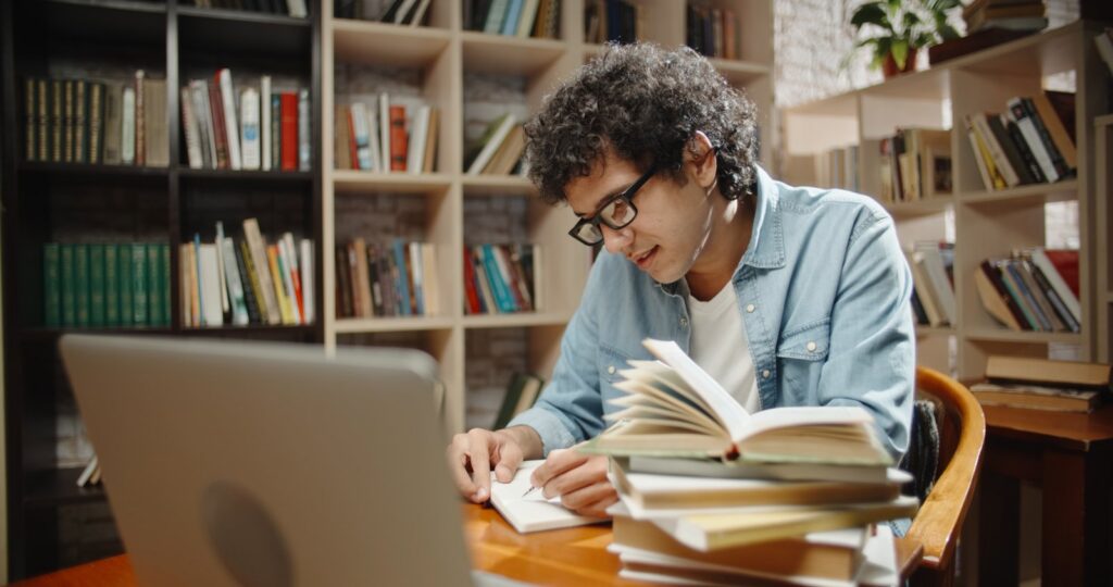 A young man in glasses takes notes at a laptop surrounded by stacks of open books in a home library
