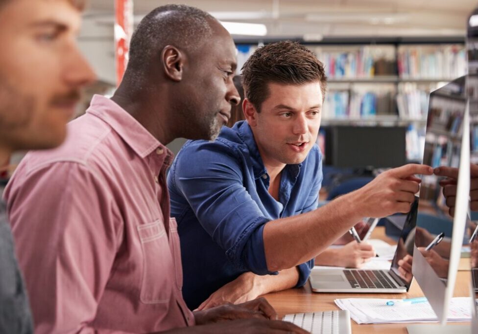 A young man points at a screen to help two adult learners at computers in a library lab