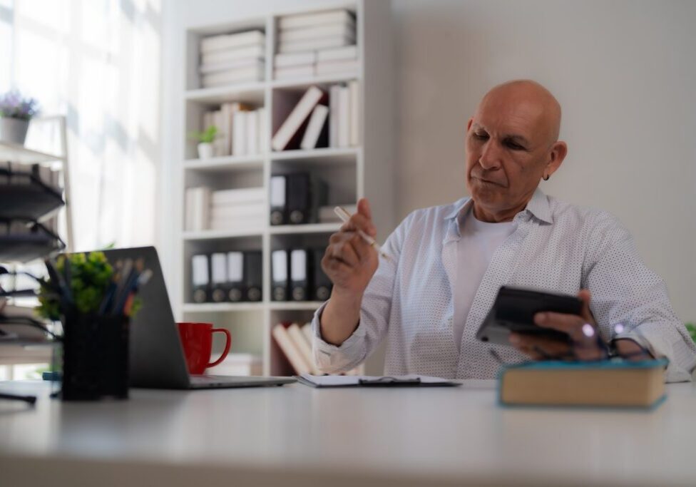 An older man in a light shirt works with a calculator and notebook beside a laptop at a bright desk