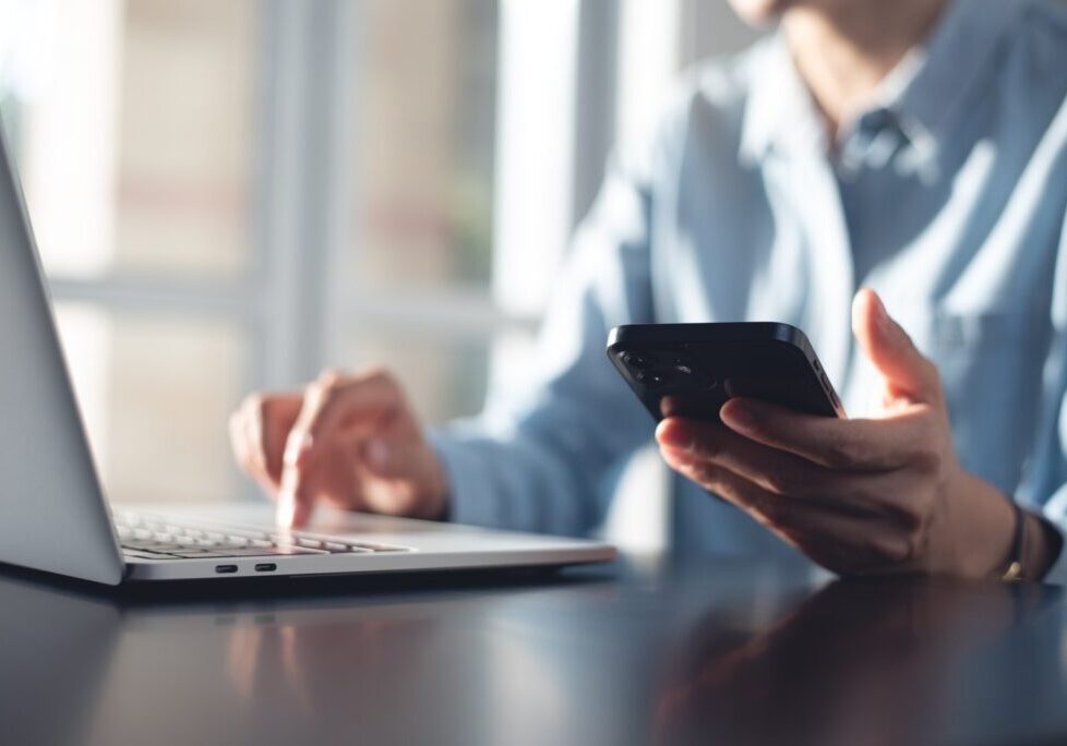 Close-up of a person using a smartphone in one hand while typing on a laptop with the other
