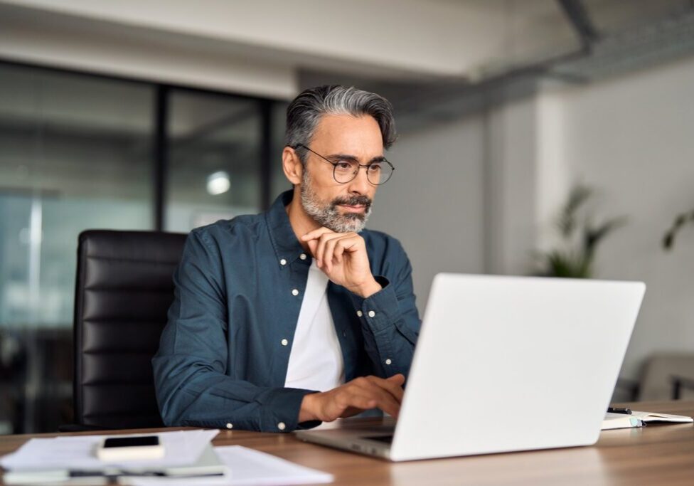 A grey-haired man in a navy shirt rests his chin on his hand while reading his laptop screen
