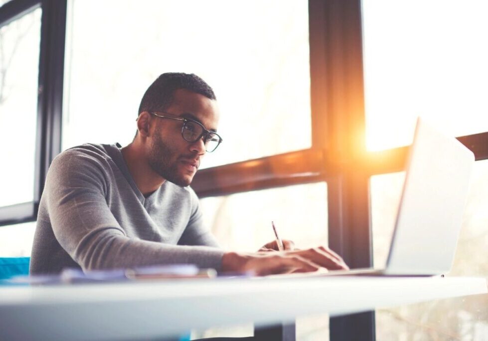 A man writing on paper with a laptop nearby in bright sunlight.