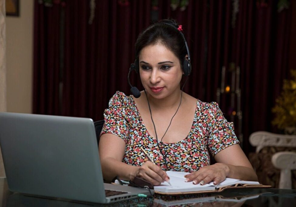 A woman wearing headphones working on a laptop and taking notes.