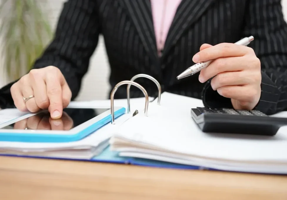 Person using a calculator and tablet with open binder on desk.