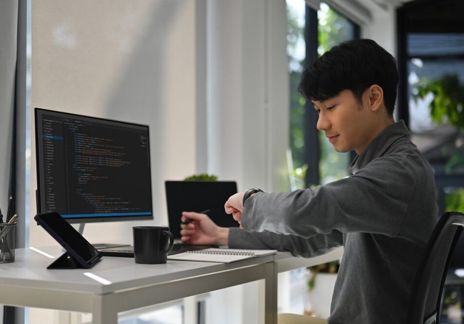 Person at desk checking wristwatch beside computer