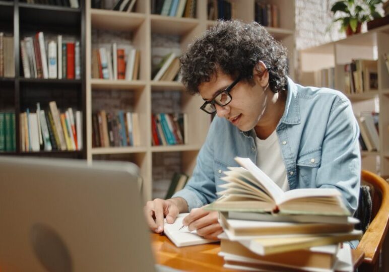 A young man in glasses takes notes at a laptop surrounded by stacks of open books in a home library
